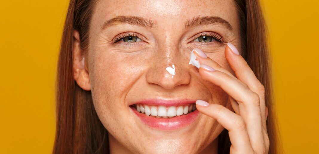smiling white woman with freckles applying sunscreen spf 30 spf 50 to her face on a yellow background
