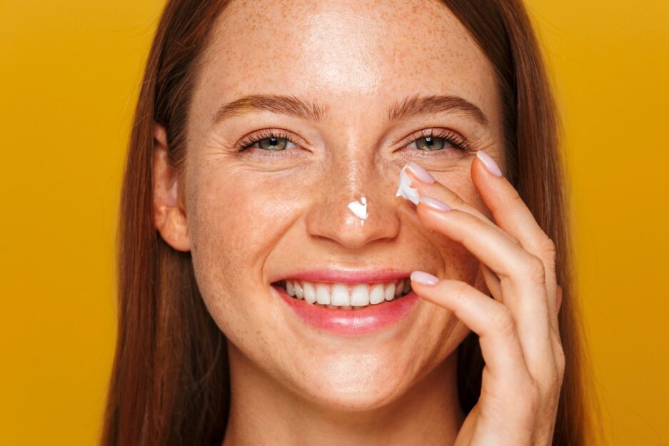 smiling white woman with freckles applying sunscreen spf 30 spf 50 to her face on a yellow background